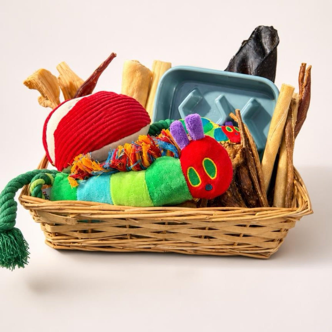 Basket of dog toys including a colorful caterpillar toy, natural bones, and a chew block on a white background